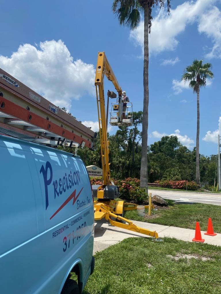 A worker in a bucket lift attached to a Precision Electric van works on a palm tree outdoors on a sunny day.