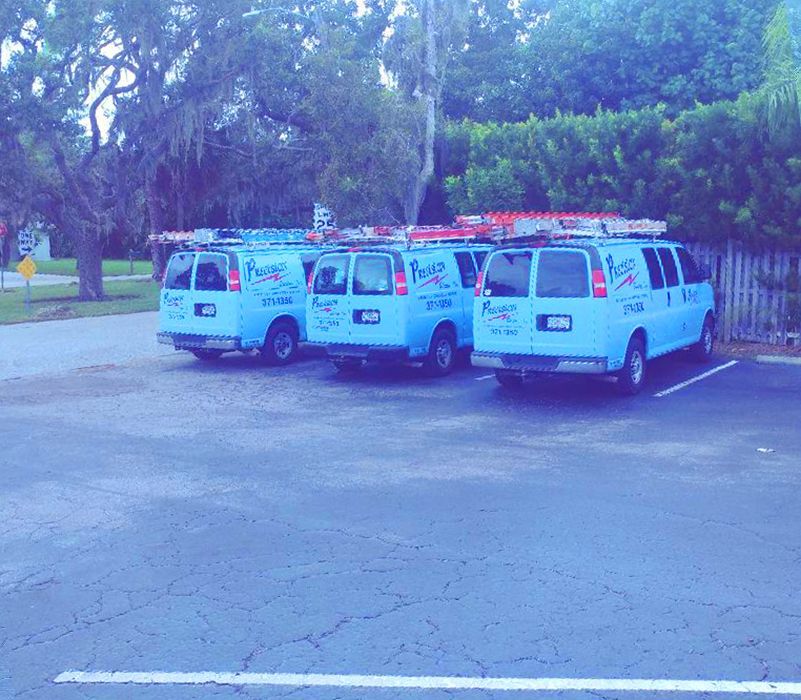 Three light blue service vans with roof ladders parked in a row on an asphalt lot near trees and a fence.