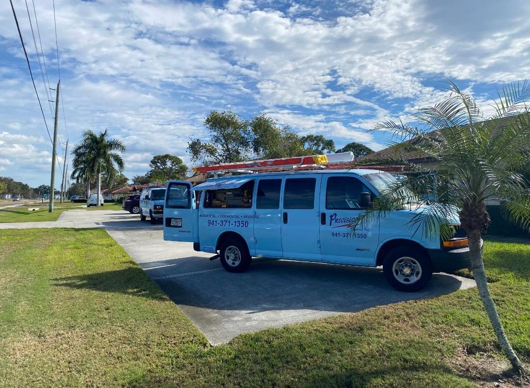 A light blue work van with a roof ladder rack parked on a paved driveway in a sunny, grassy area with palm trees.