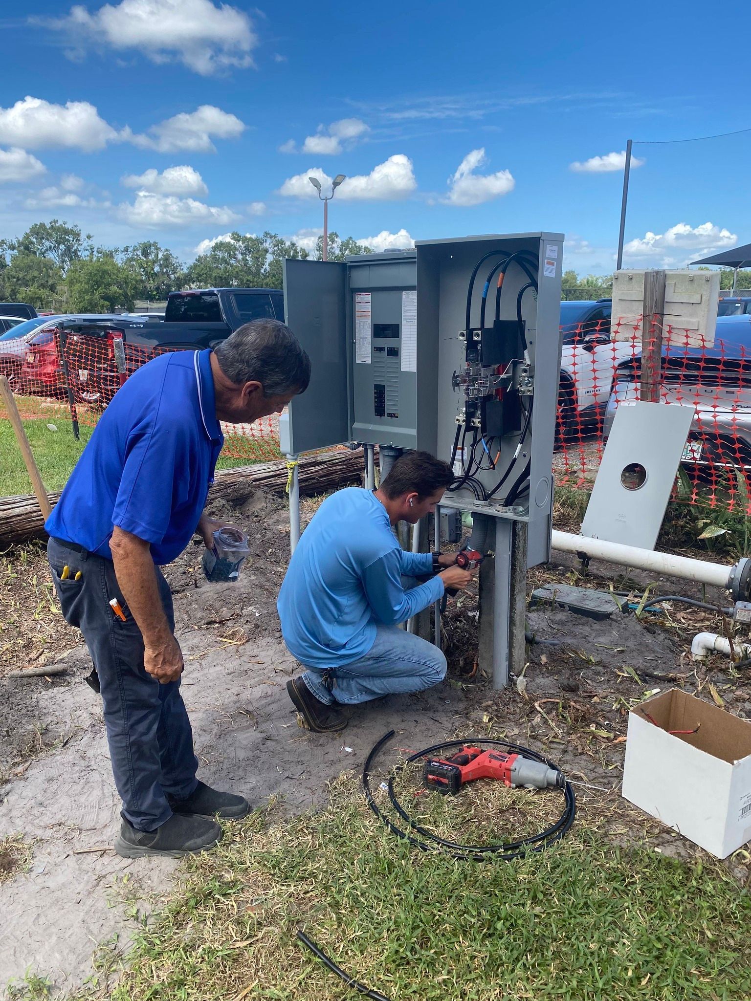 Two workers inspect electrical equipment beside a utility box outdoors.