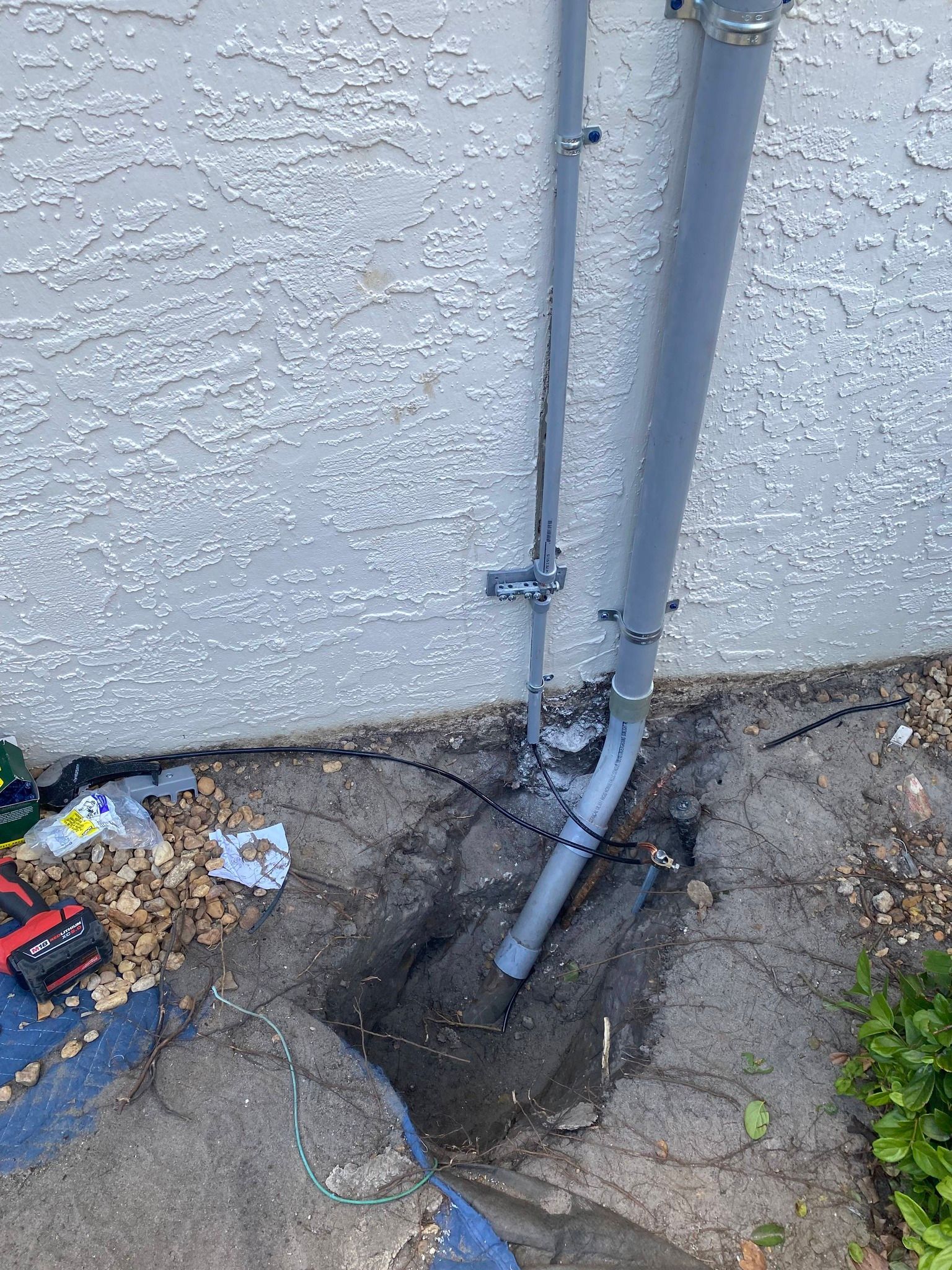 Damaged utility pipe at wall base with exposed cables and debris on dirt ground outdoors.