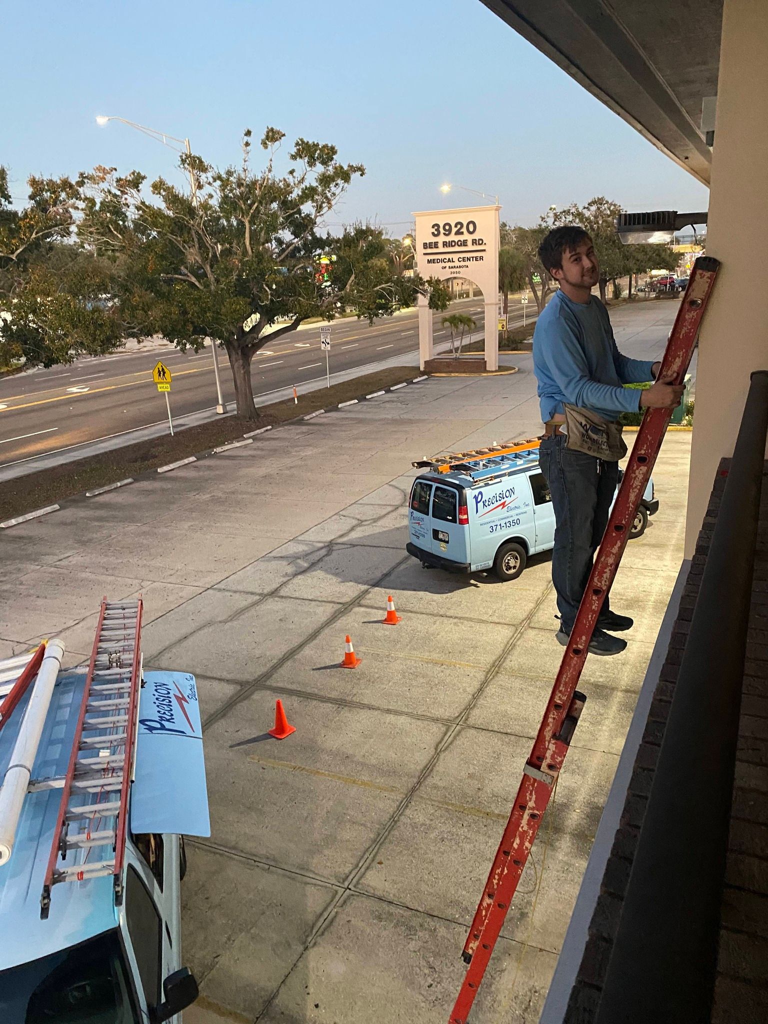 Worker on ladder beside an airport curb with orange cones and a blue utility cart at sunset.