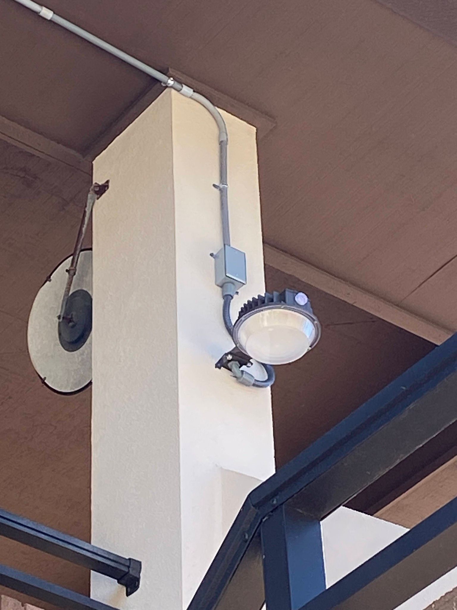 Outdoor wall-mounted security light with a white fixture on a beige column under a brown roof.
