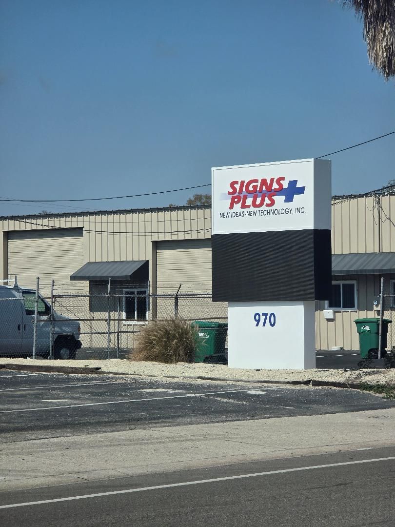 Industrial building with a “Signs Plus” sign, fenced lot, and white address marker 970 by the road