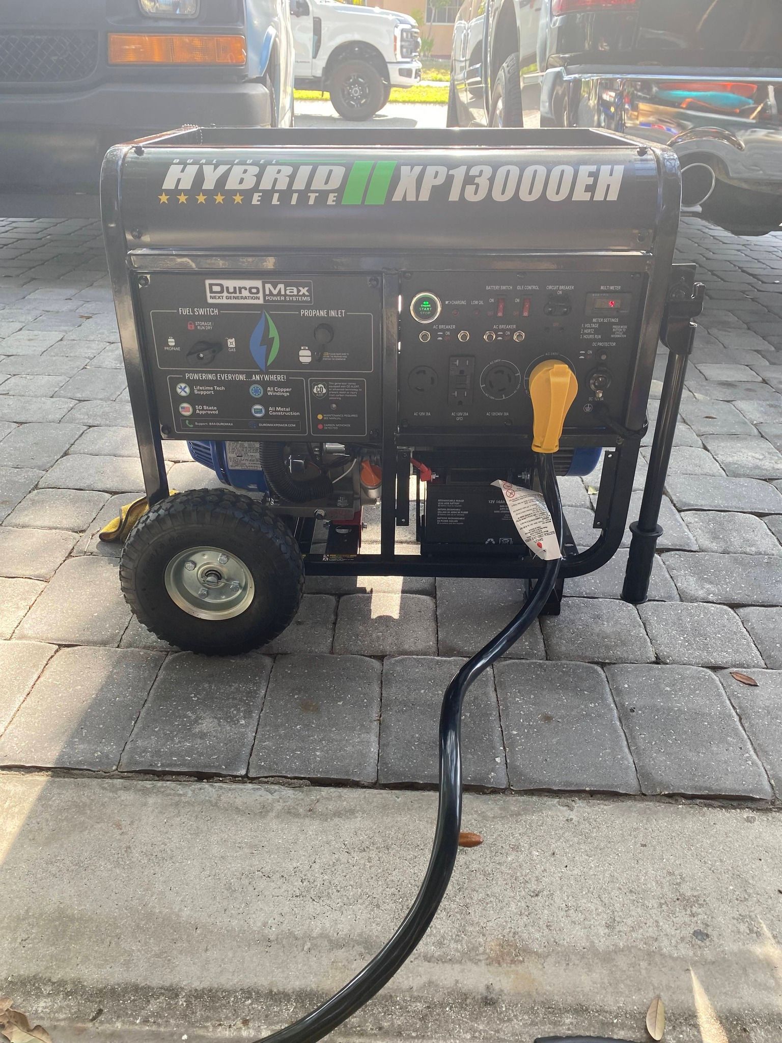 Black portable generator on a paved patio with a hose attached, outdoors near a vehicle.