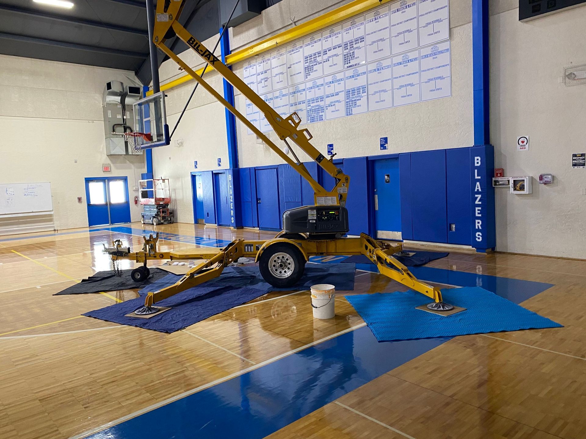 Yellow scissor lift in a gymnasium, with blue safety mats on the floor and wall padding behind it.