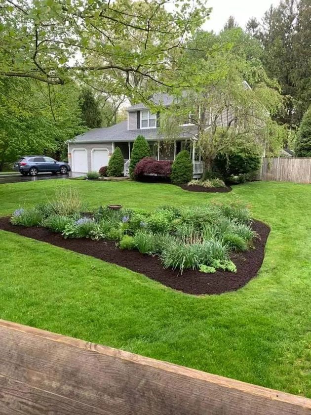 Suburban house with a white garage, green lawn, and landscaped flower bed under trees