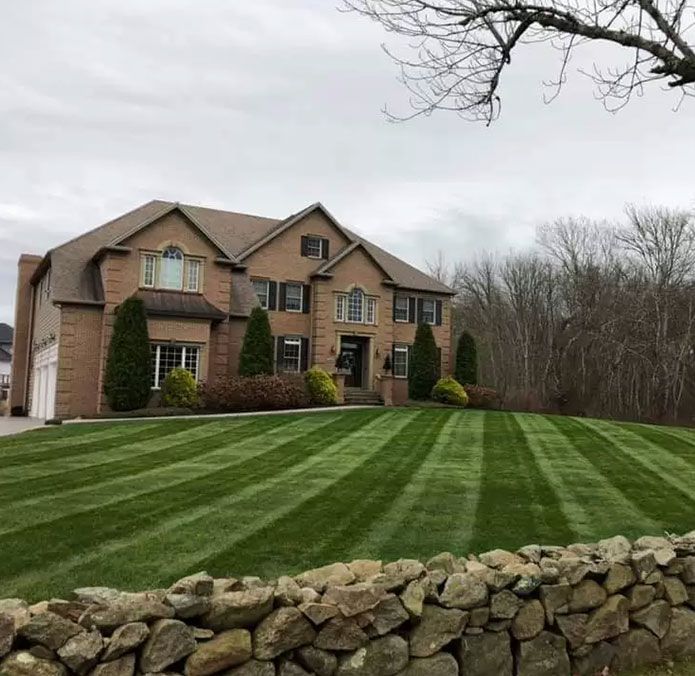 Large brick house with striped green lawn and stone wall under a cloudy sky