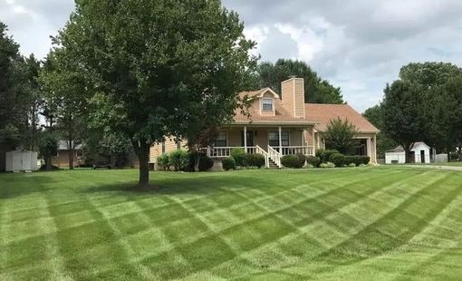 Large suburban house with a covered porch, surrounded by a striped green lawn and trees under cloudy skies