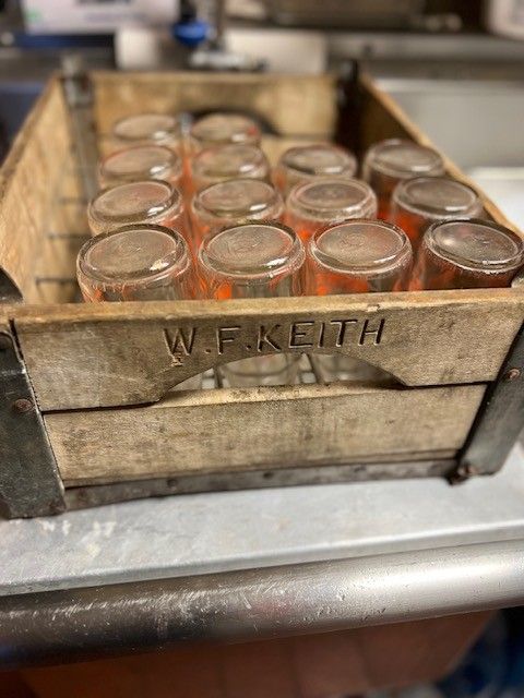 Wooden crate holding a grid of clear glass jars, labeled with