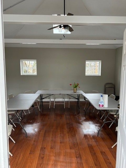 An empty room with tables arranged in a U-shape. Hardwood floor, gray walls, and two small windows.