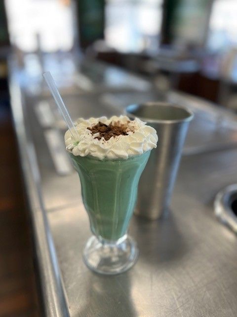 Green milkshake topped with whipped cream and chocolate shavings, with a metal shaker on a bar.