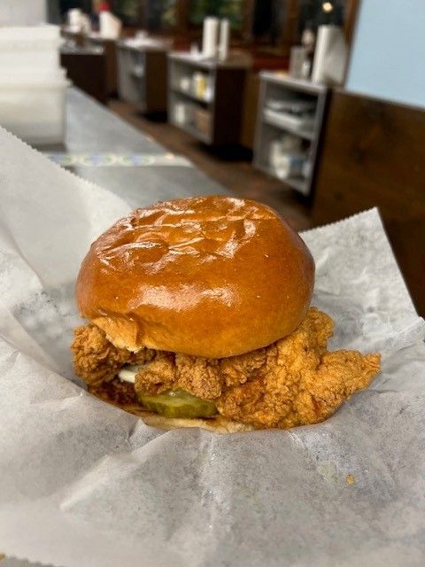 Fried chicken sandwich on a brioche bun with pickles, on white paper; kitchen in background.