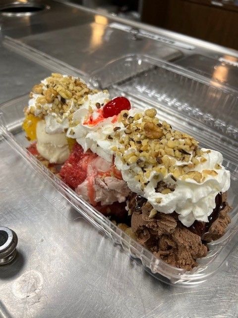 A sundae with vanilla, strawberry, and chocolate ice cream topped with whipped cream, nuts, and a cherry in a clear plastic container.