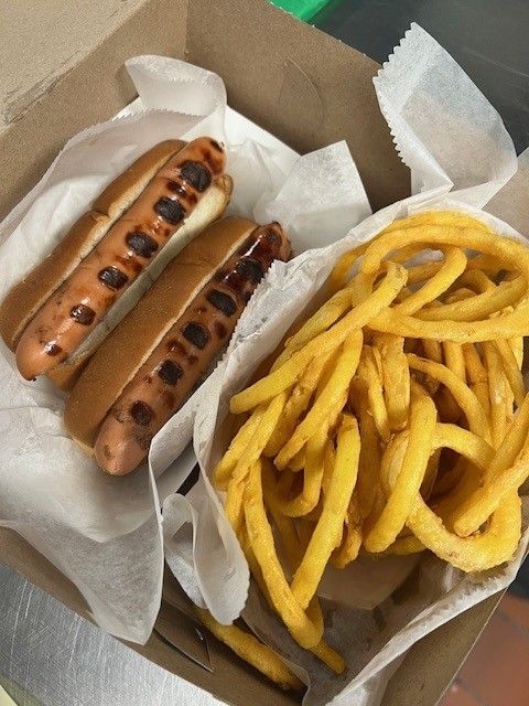 Two hot dogs with grilled marks and curly fries in a cardboard container.