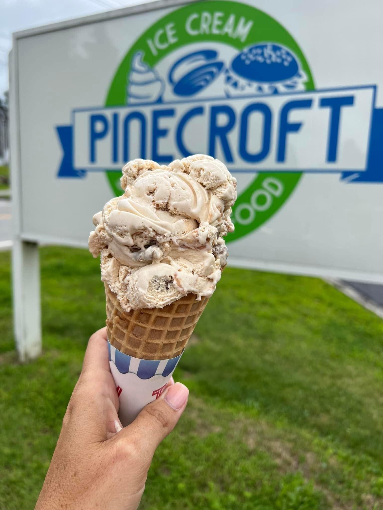 Person holding ice cream cone in front of a Pinecroft Ice Cream sign.
