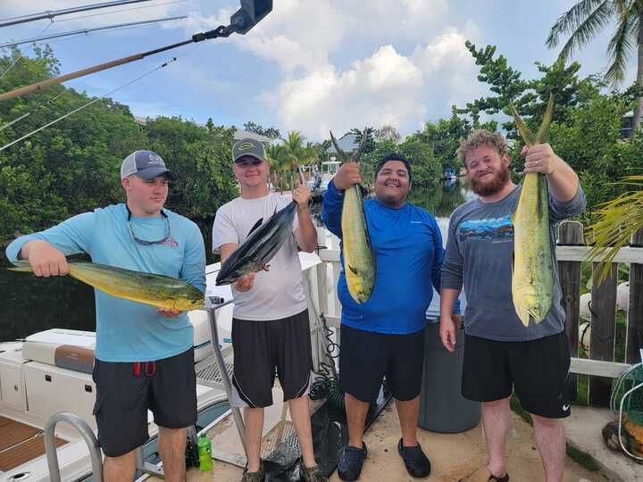 Four people on a boat holding up fish they caught. Brightly colored fish, blue sky, green trees.