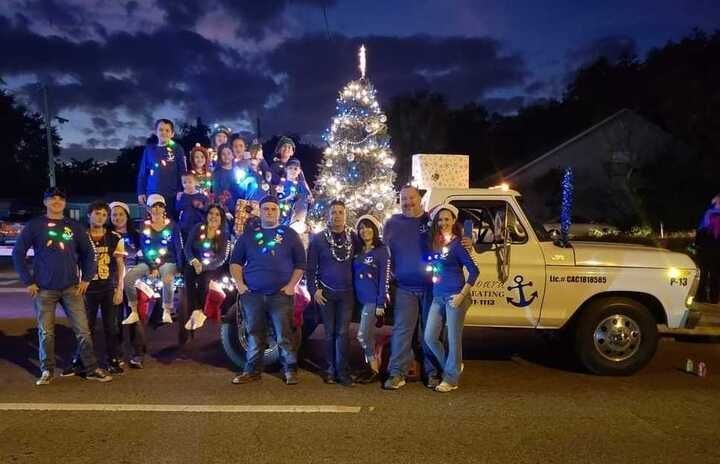 Group of people in blue sweaters pose with a decorated truck and Christmas tree at night.