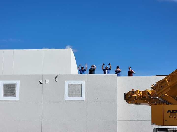 People waving from a white building's roof with a yellow crane in the foreground against a blue sky.