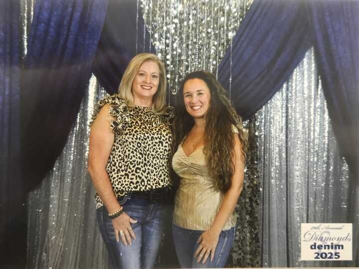 Two women posing in front of a blue and silver backdrop. One wears a leopard print top, the other a gold top, both with jeans.