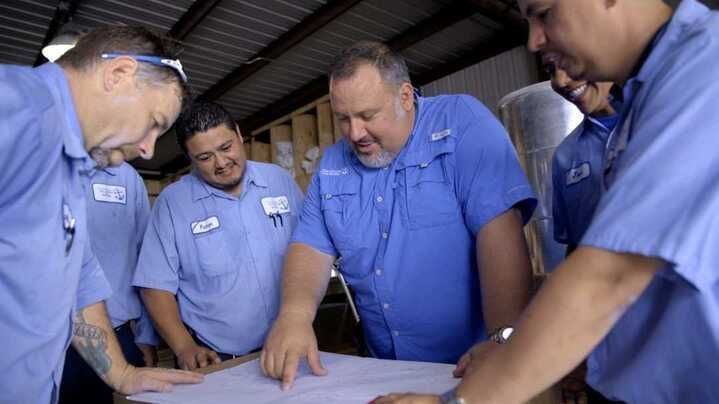Five people in blue shirts looking at blueprints in a warehouse.