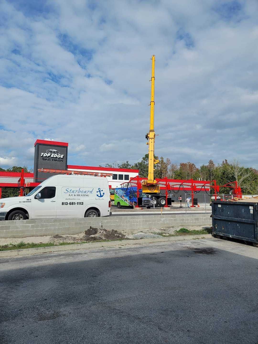 Crane at a construction site near a building and a parked van. The sky is cloudy and grey.