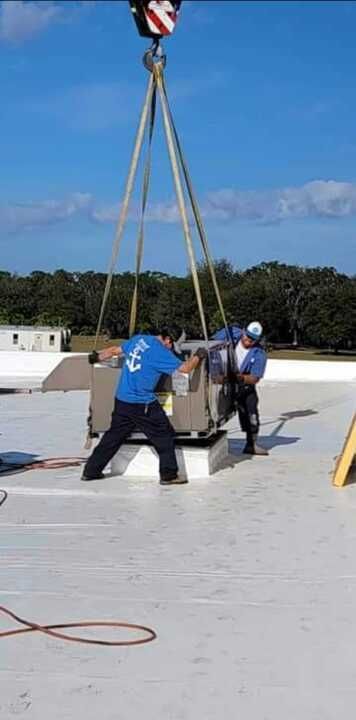 Workers installing HVAC unit on roof using a crane on a sunny day.