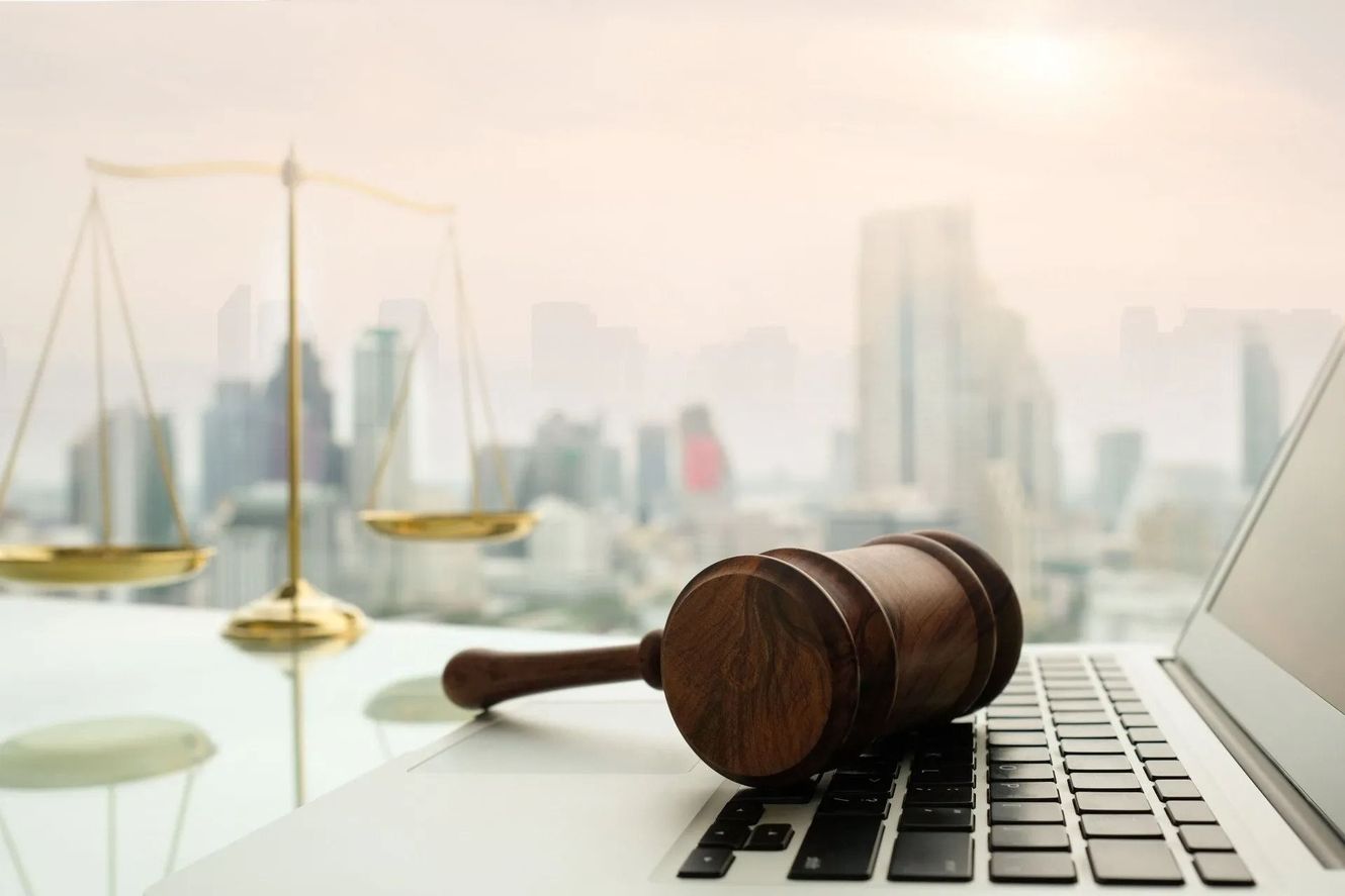 Gavel on laptop keyboard next to scales of justice, cityscape in the background.