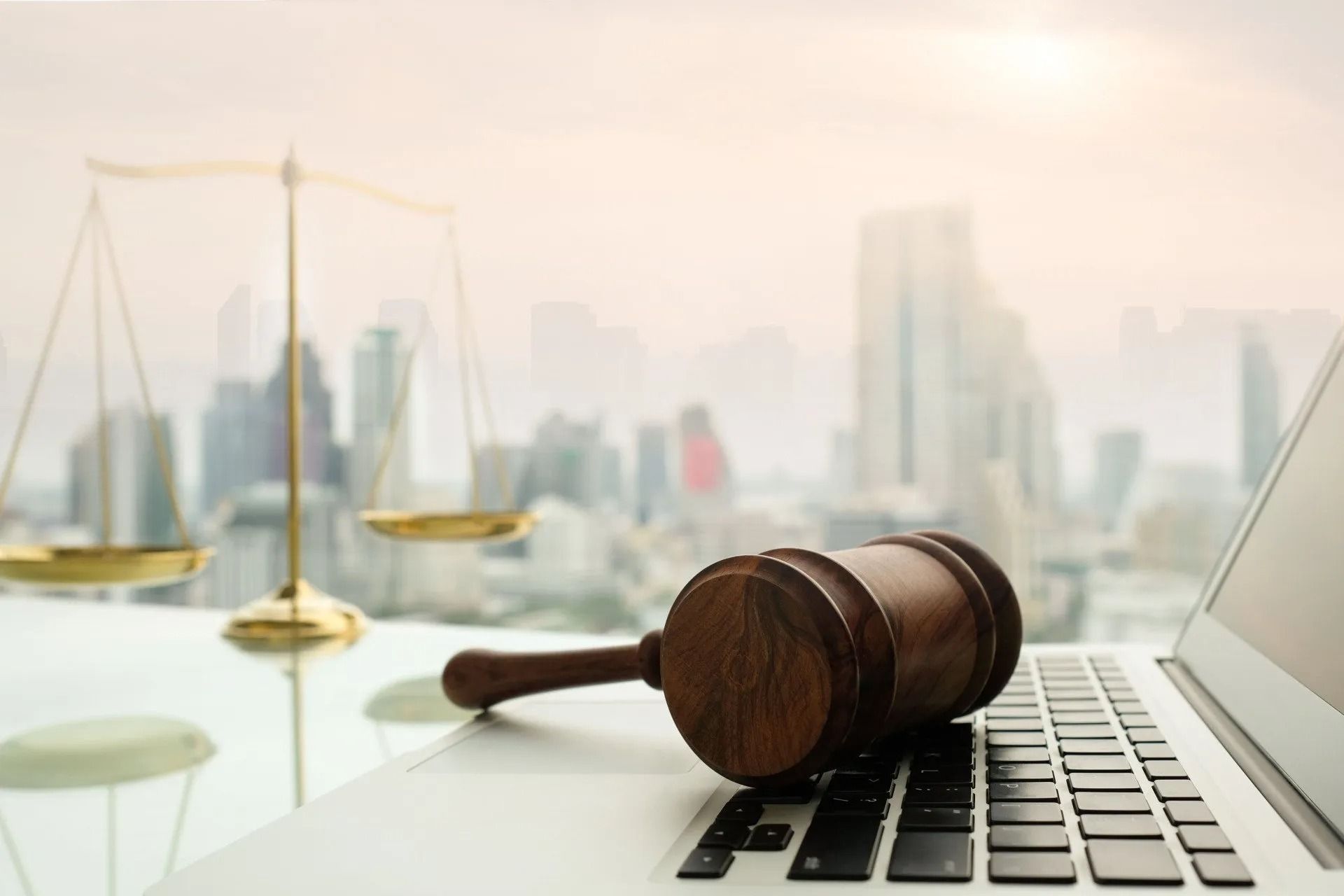 Gavel on laptop keyboard next to scales of justice, cityscape in the background.