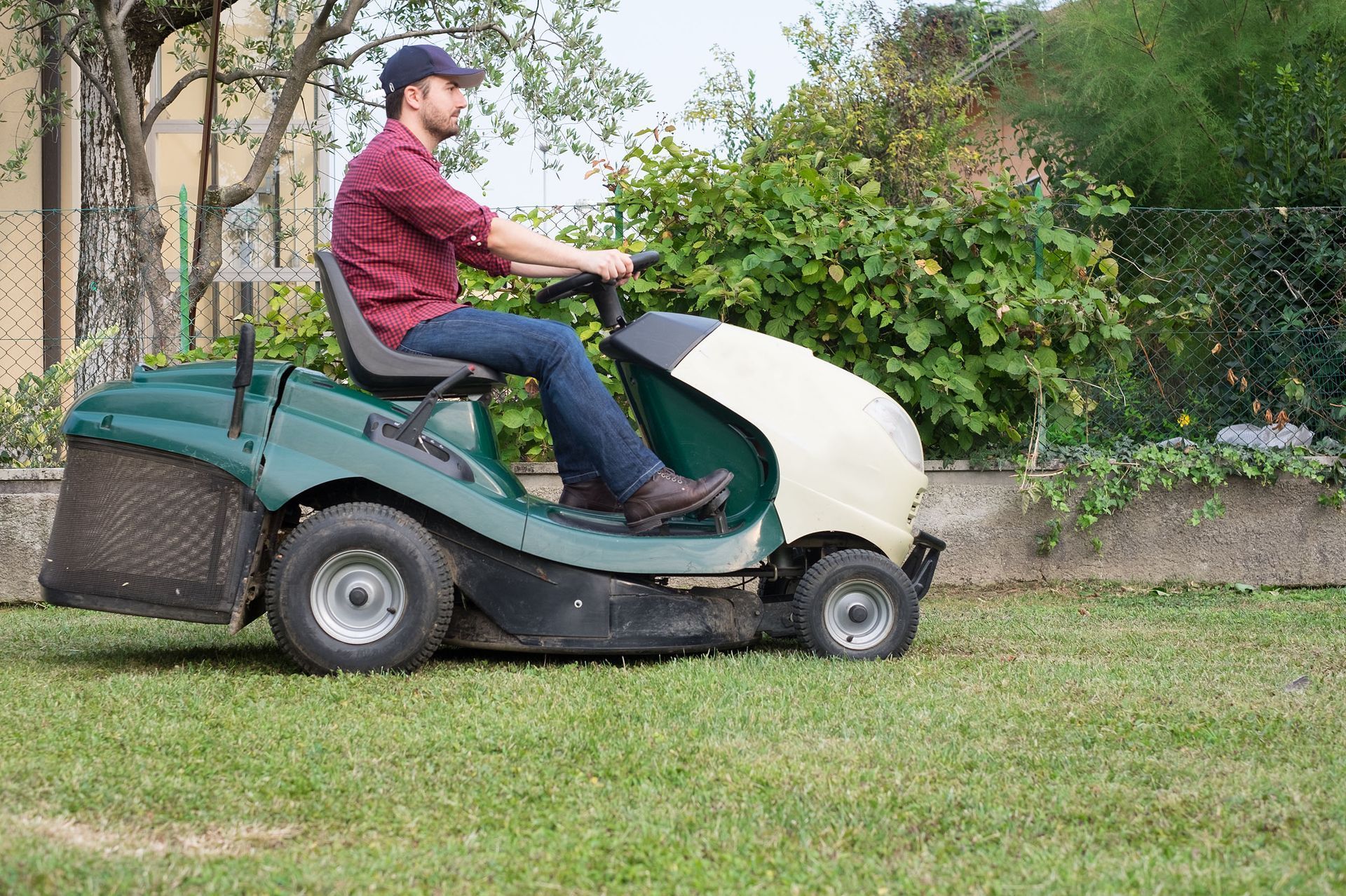Man driving a ride-on mower across a grassy yard with trees and bushes in the background