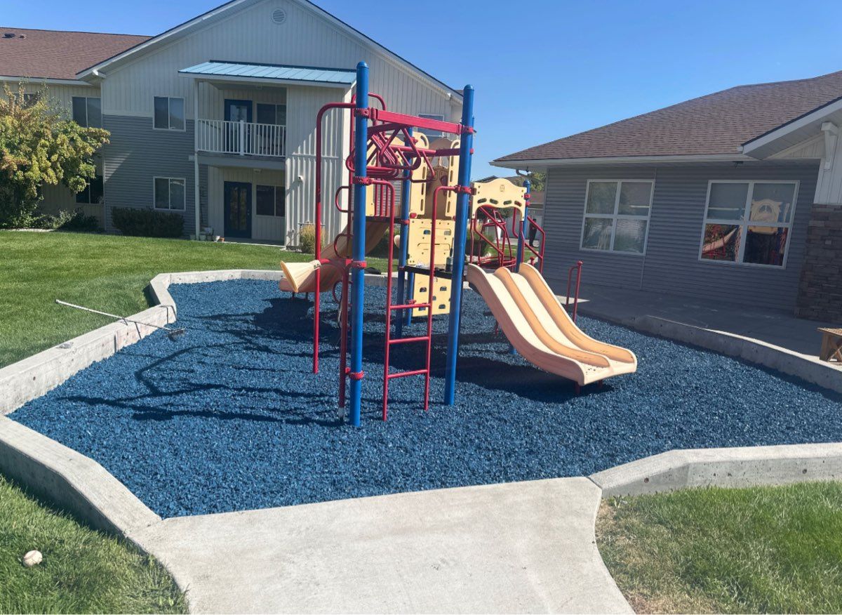 Colorful playground structure on blue mulch in a residential backyard between two houses.