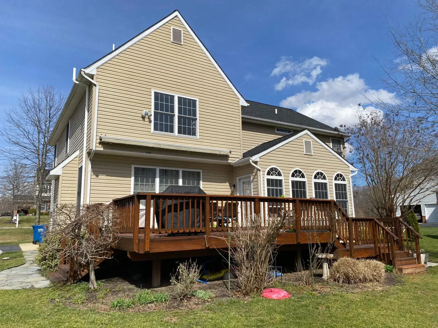 Two-story beige house with a wooden deck and stone accents under a blue sky