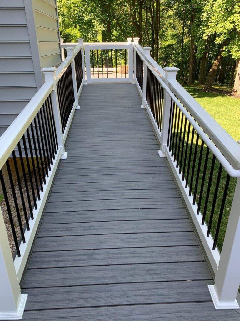 White-railed wooden ramp leading from a house to a yard, with gray deck boards and trees beyond