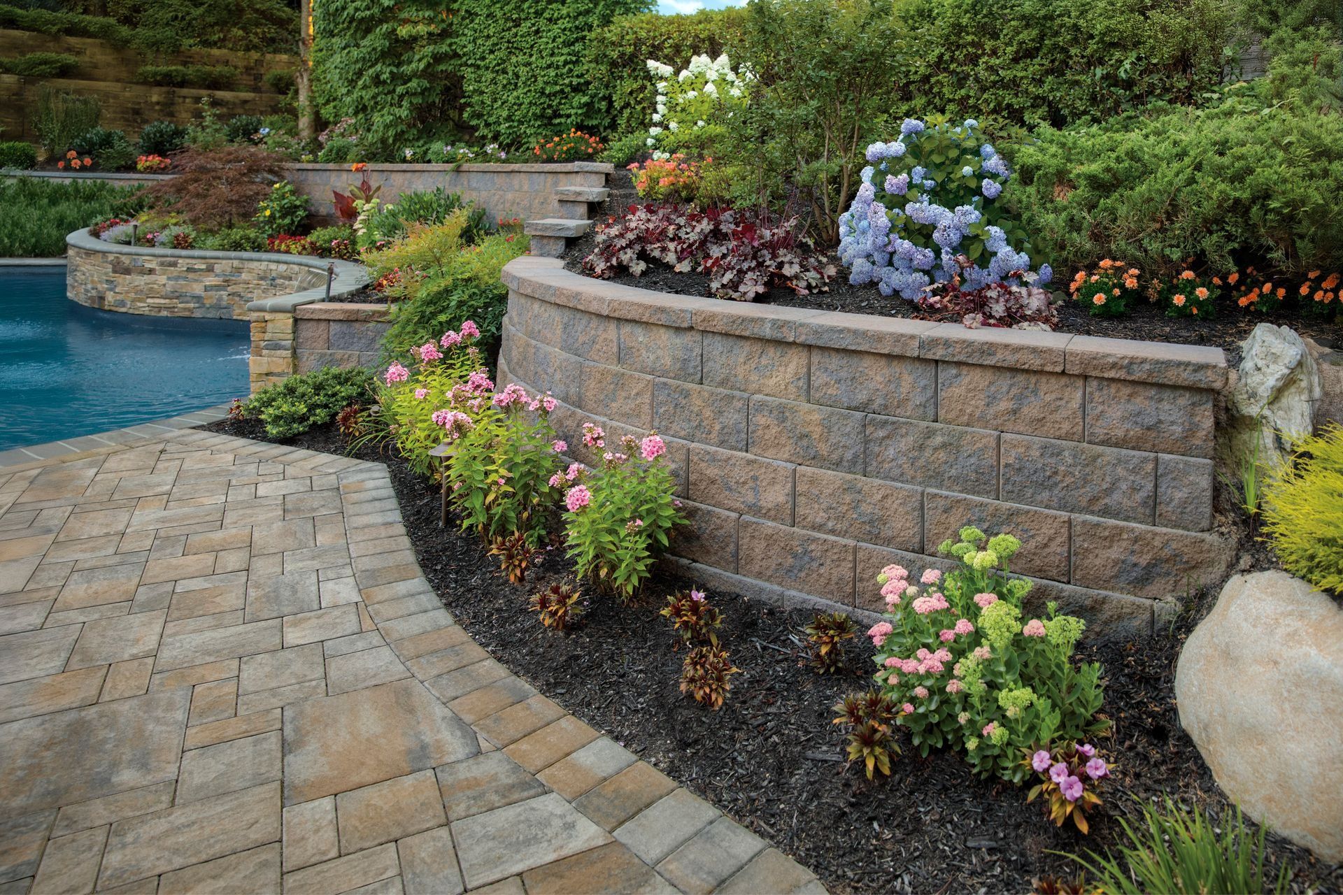 Curved stone patio beside a blue pool and raised flower bed with pink and purple blooms