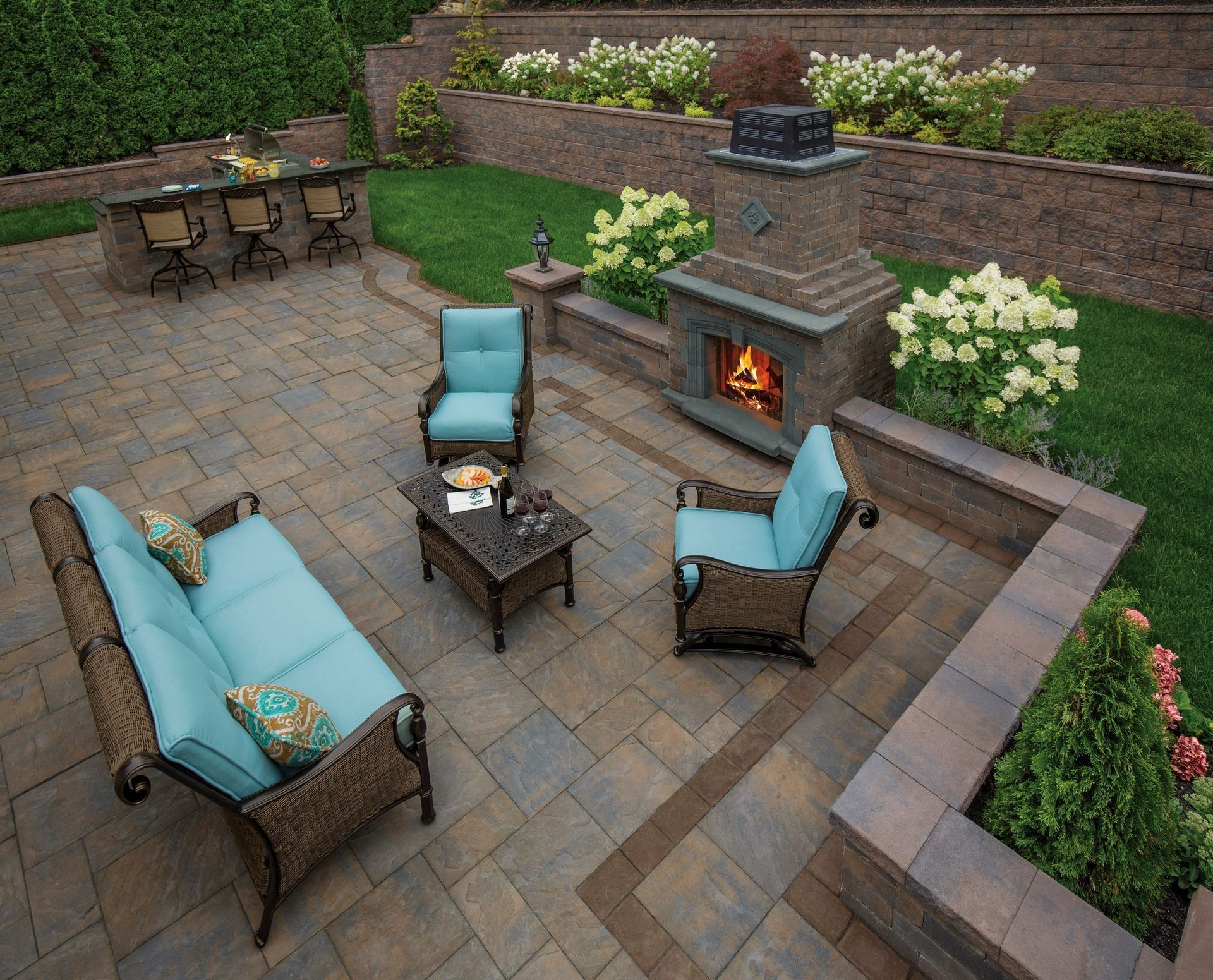 Patio lounge with blue chairs around a fire pit and stone outdoor fireplace, surrounded by greenery and flowers