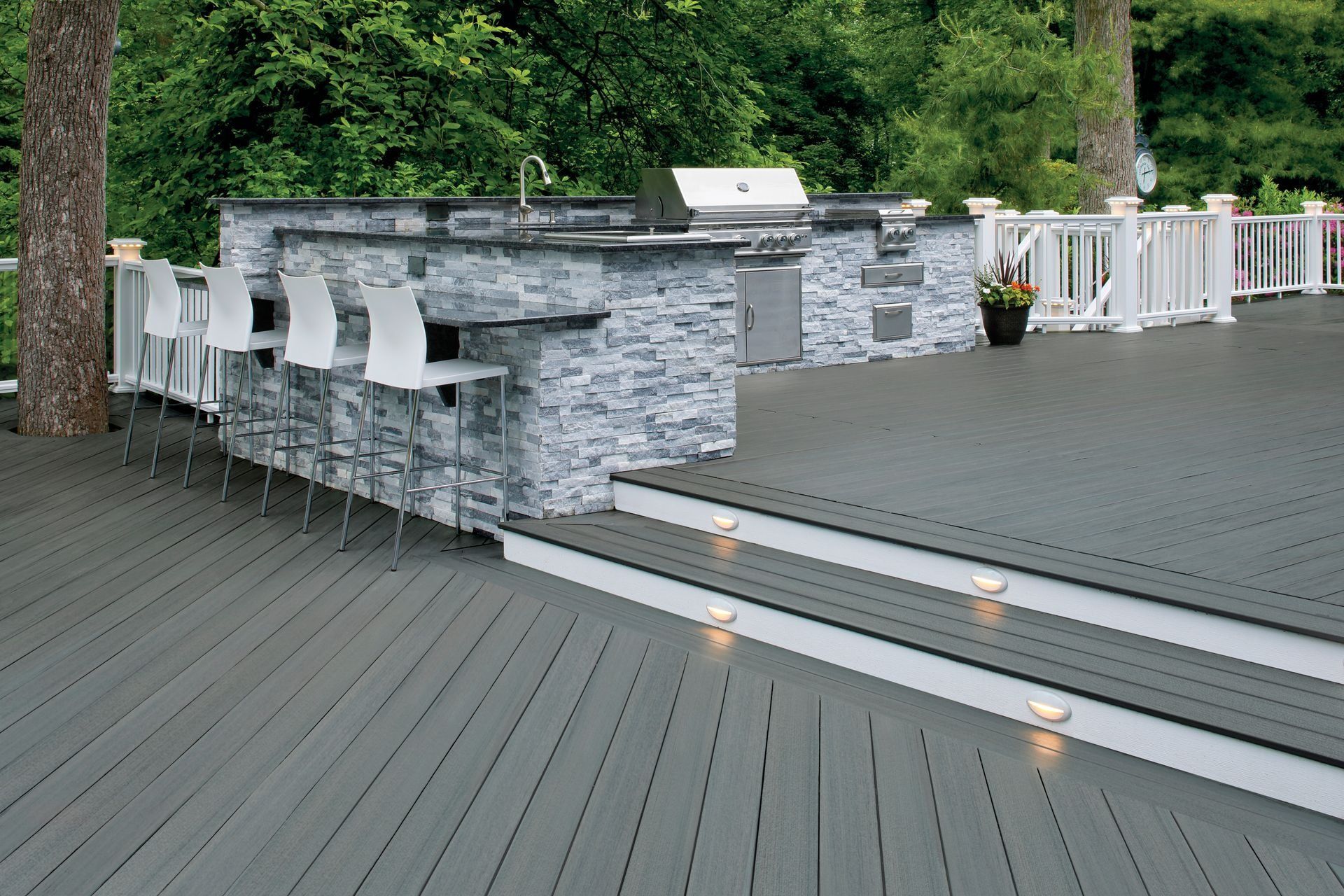 Stone outdoor patio with white chairs, gray deck flooring, and warm step lights beside greenery