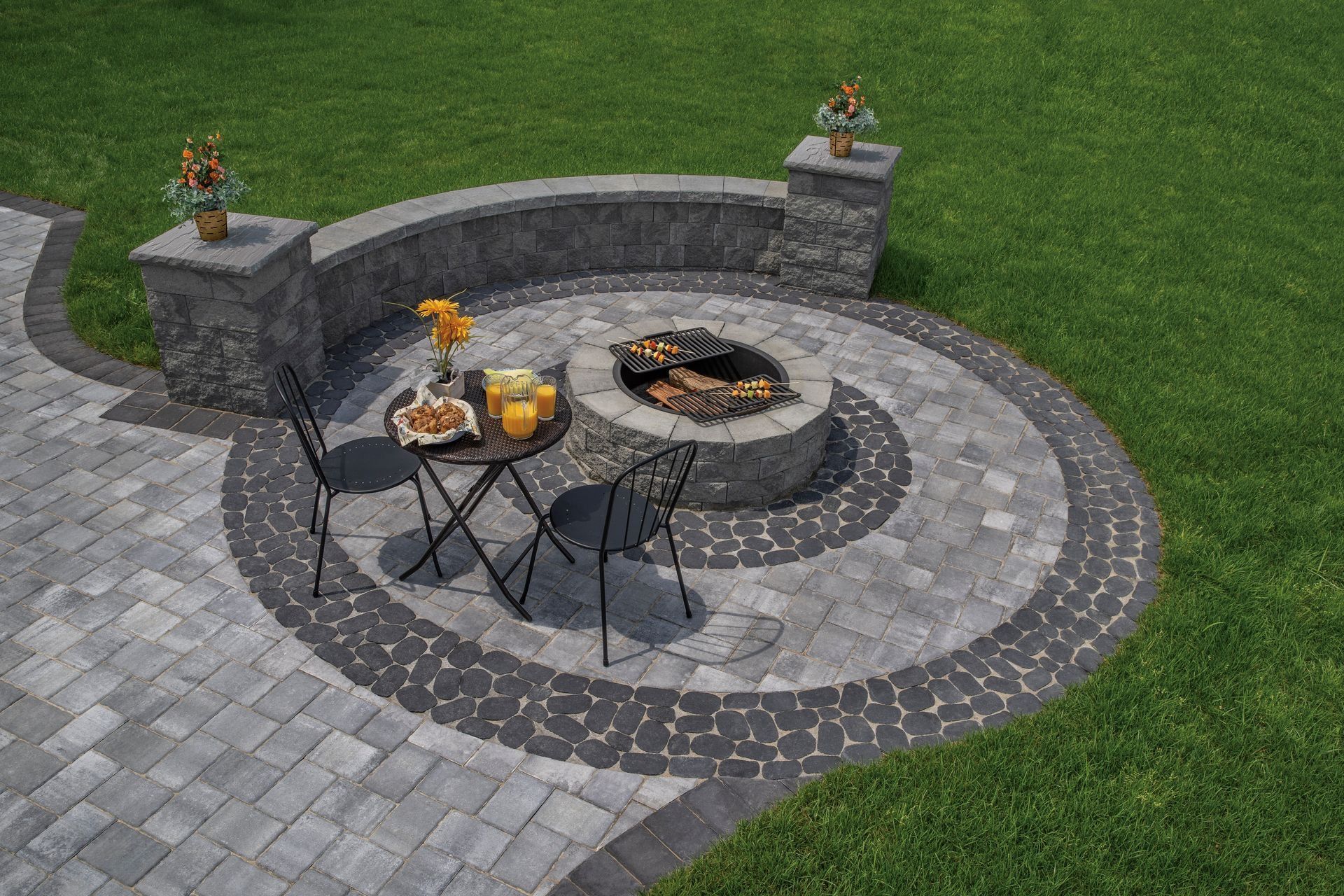 Stone patio fire pit with round seating area, two chairs, and small side tables on a grassy lawn.