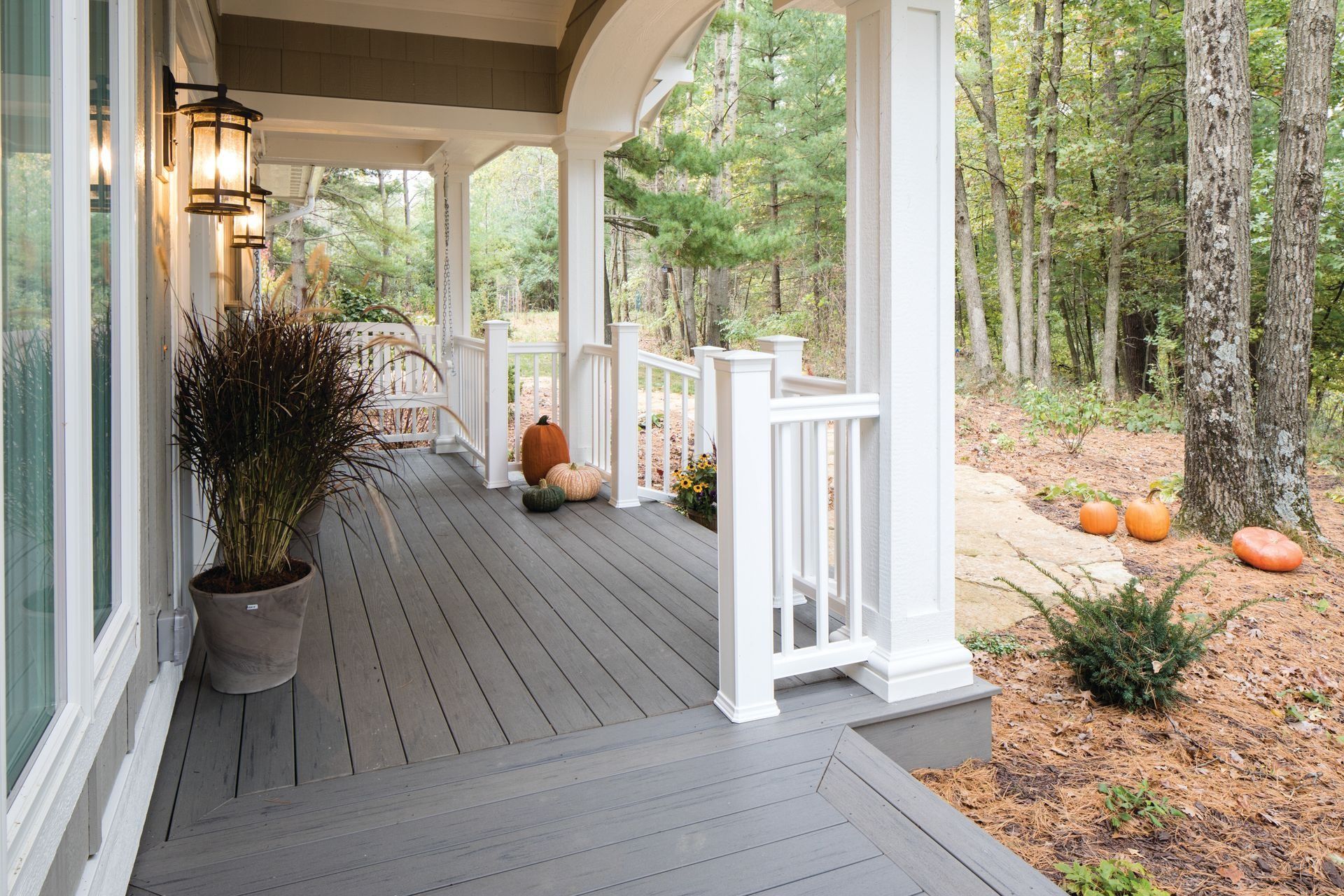 Covered gray porch with pumpkins, potted plants, and white railings overlooking a wooded yard