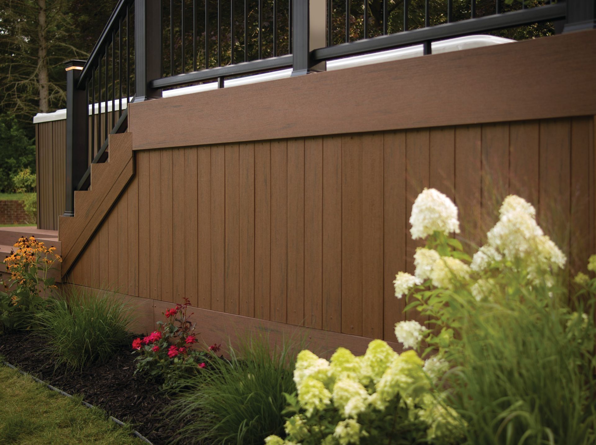 Brown wooden retaining wall beside a garden with white and red flowers and black railing above