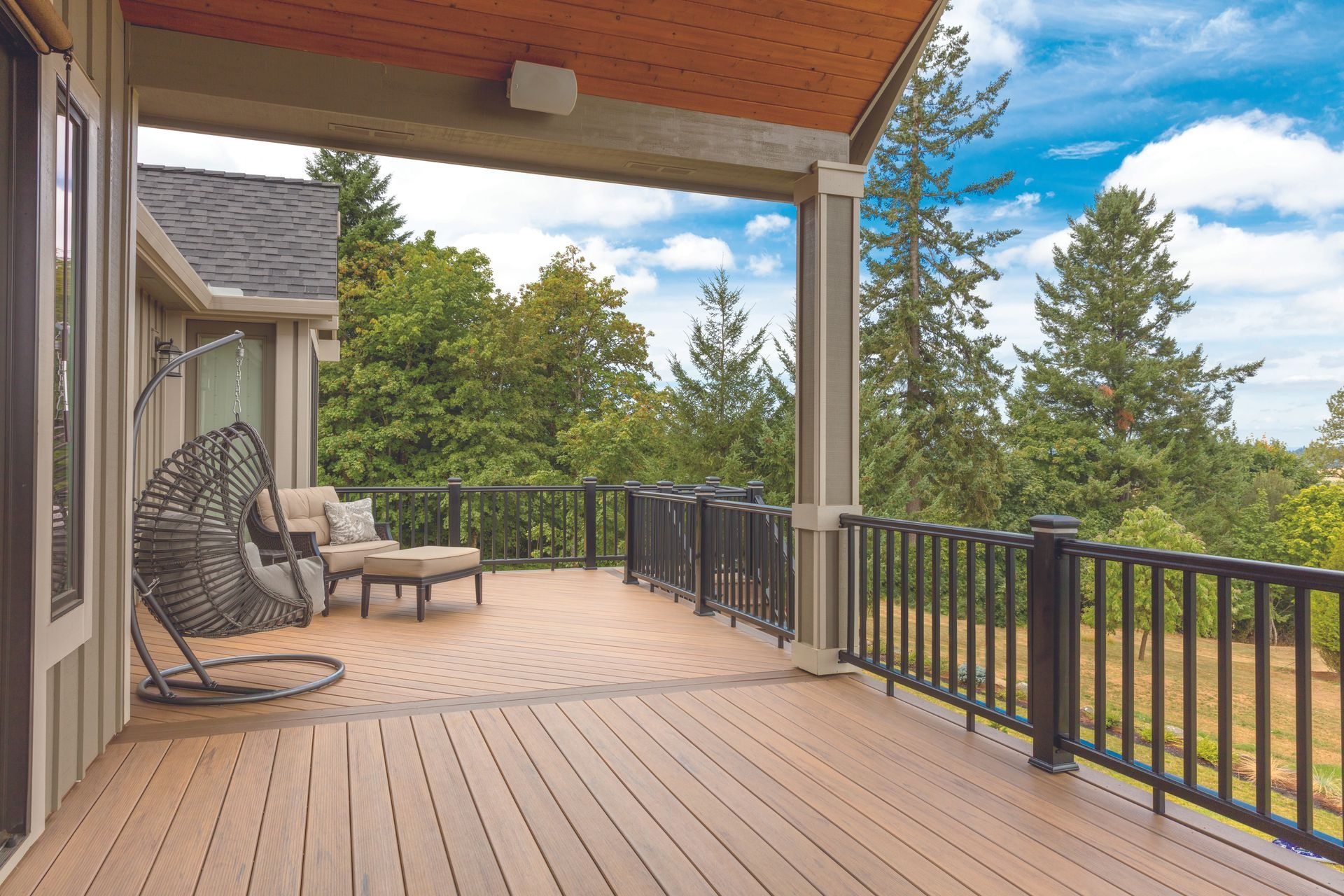 Spacious wooden deck with black railing, cushioned swing chair, and forested backyard view