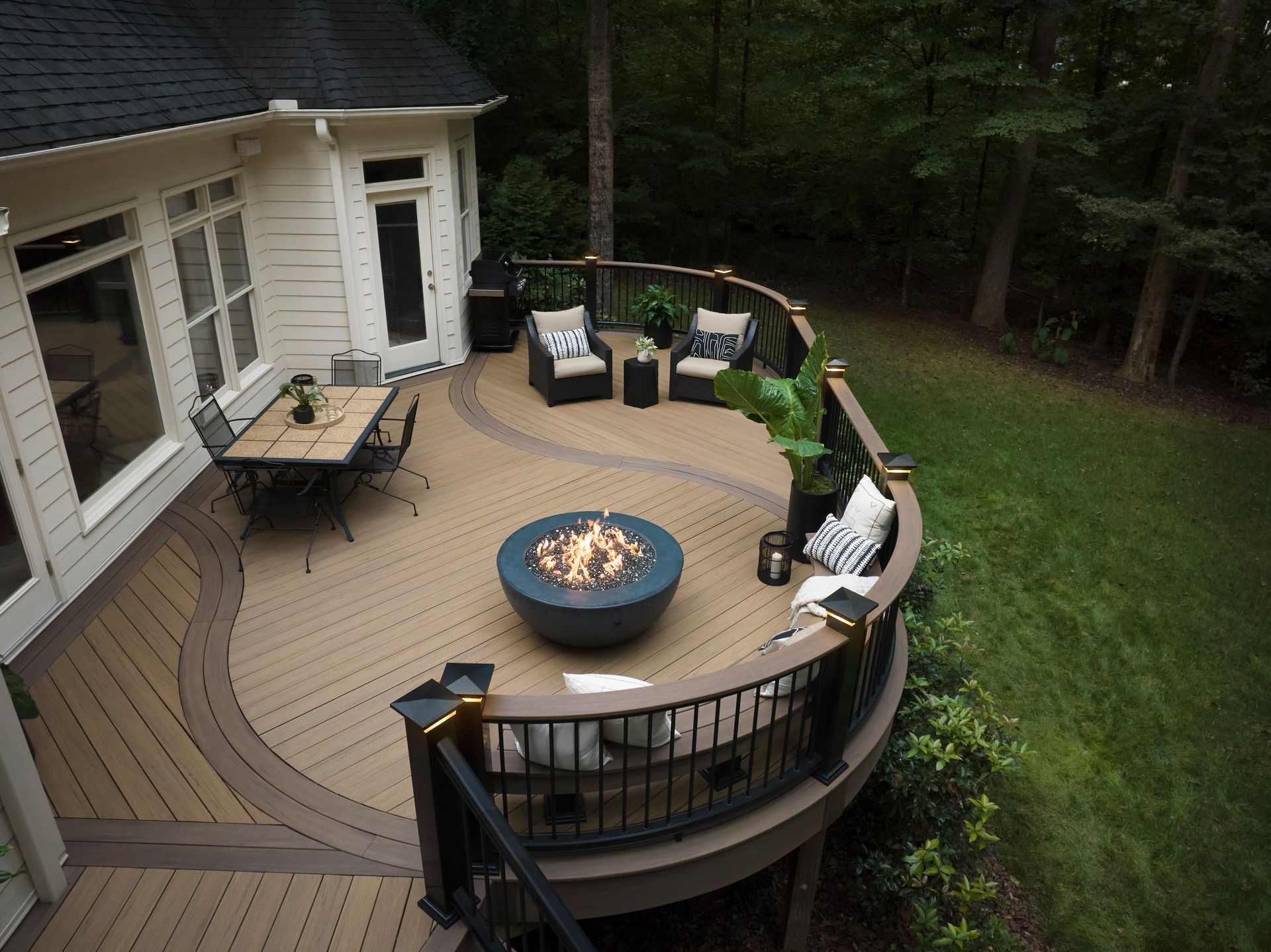 Curved backyard deck with a fire pit, outdoor chairs, and a table beside a wooded yard.