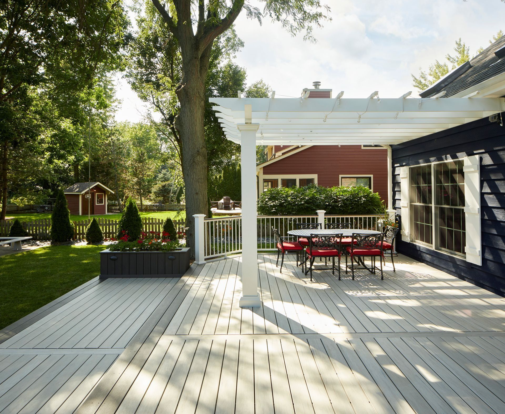 Wooden backyard deck with red outdoor table and chairs under a white pergola beside a house.