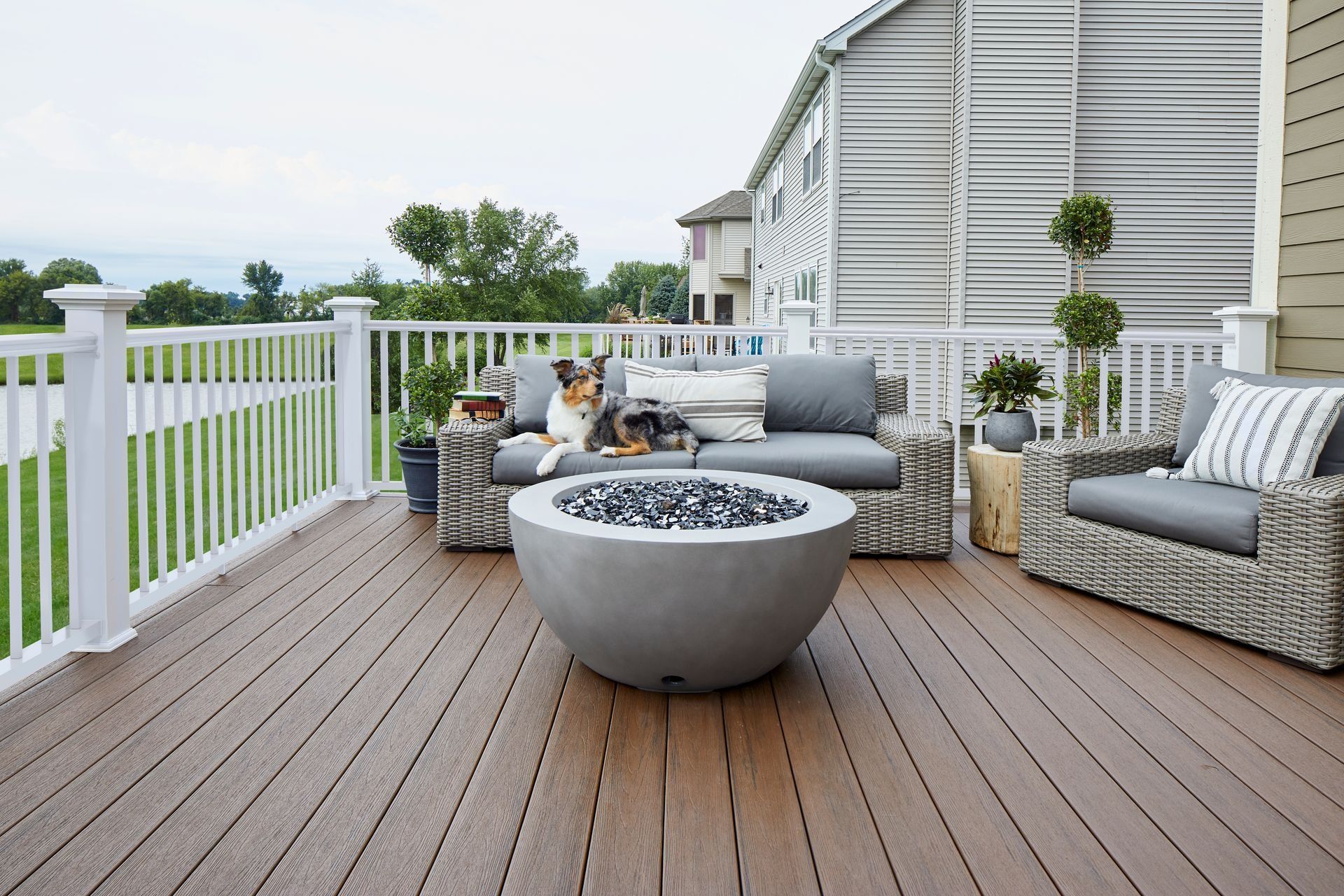 Patio deck with gray seating and a round fire pit beside a white railing and house exterior