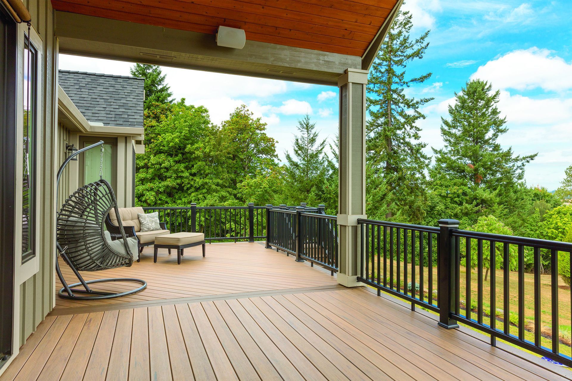 Covered wooden deck with black railing, chair swing, and green trees overlooking a yard.