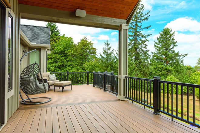 Covered wooden deck with black railing, chair swing, and green trees overlooking a yard.
