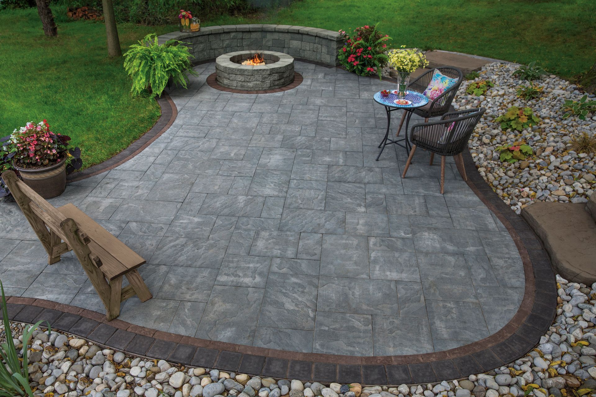 Curved stone patio with fire pit, bench, table and chairs, bordered by flowers and gravel