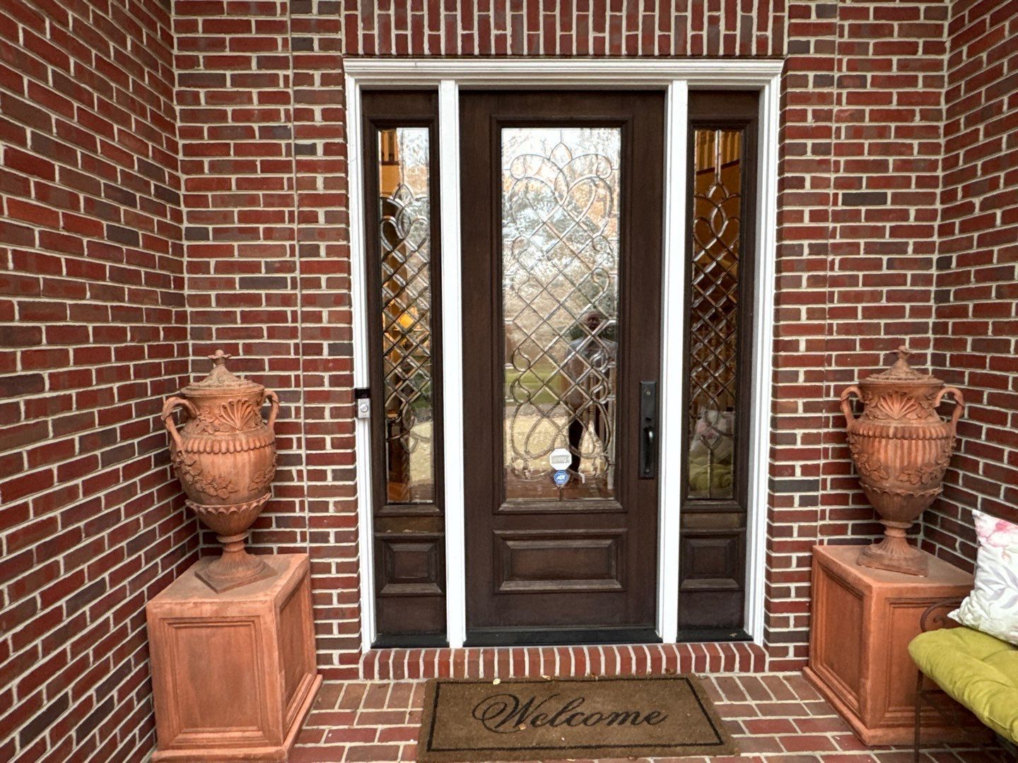 Brick entryway with double wooden doors, potted urns, and a “Welcome” mat on a front porch