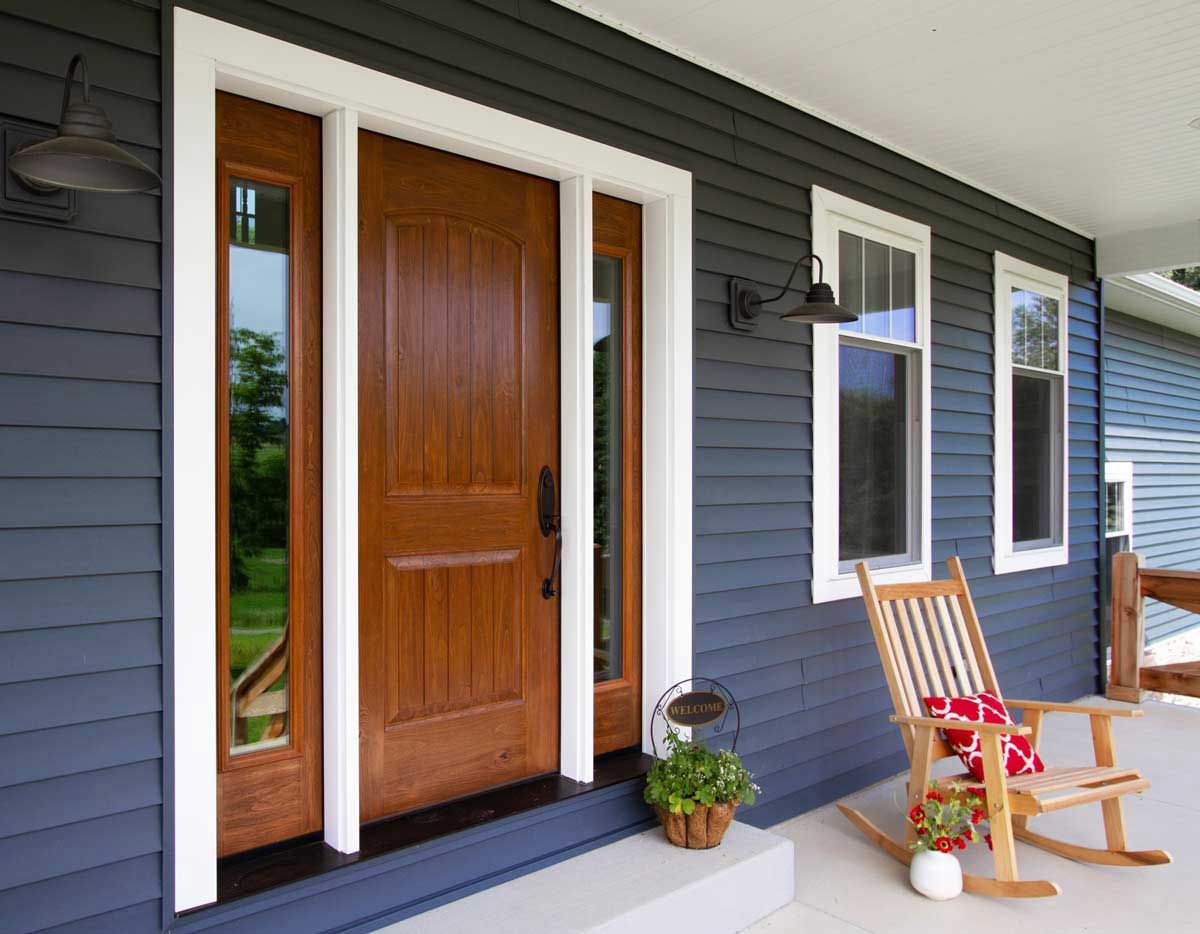 Front porch with wooden door, gray siding, white trim, and a rocking chair beside potted plants.