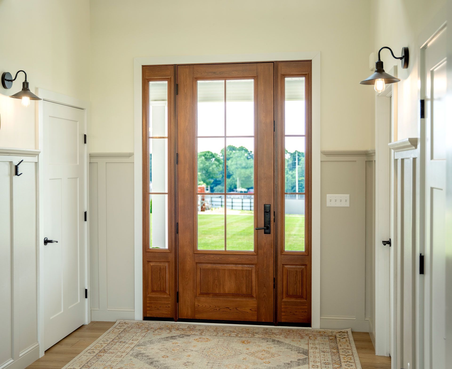 Front entry hallway with double wooden glass doors and wall sconces, showing a green lawn outside