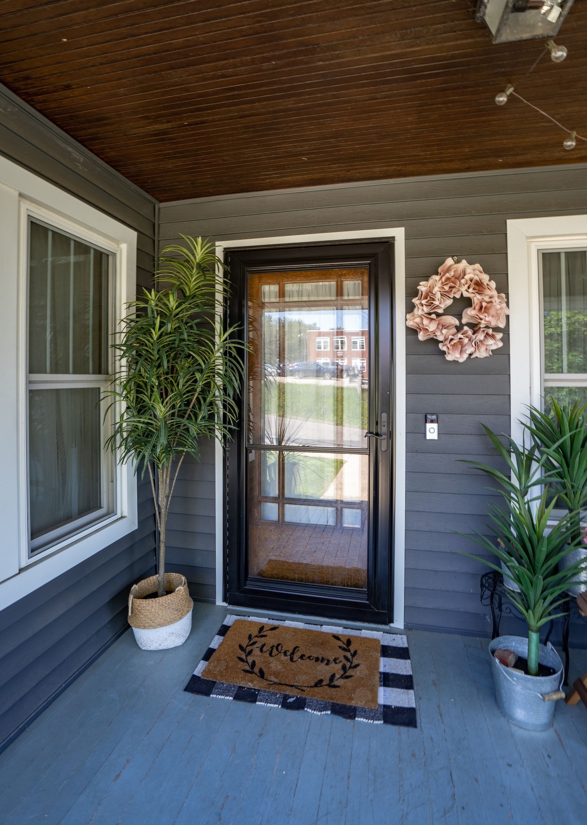 Front porch with black door, striped rug, potted plants, and gray siding beneath a wood ceiling